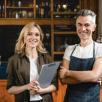 Kleinunternehmer (Barista) mit Buchprüferin, die ein Klemmbrett hält, beide stehen vor Bar-Restaurant-Café-Theke